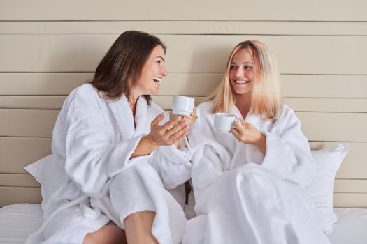 Happy mother and teenage daughter drinking coffee talking. Women in white bathrobes, resting sitting in bed, vacation together. Communication between parent and adolescent child, family, relationship