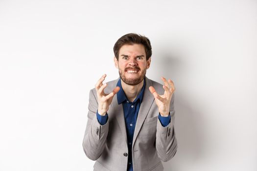 Angry and tensed businessman wants to kill someone, clenching hands and teeth, looking mad at camera, going to strangle person, standing on white background.