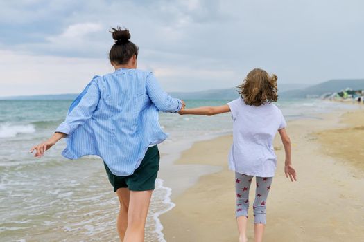 Happy mom and daughter running on sea beach holding hands, back view. Family middle-aged mother and preteen child together, vacation, sea weekend, travel, love, leisure, happiness and joy