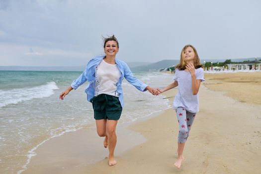 Happy mom and daughter running on sea beach holding hands. Family middle-aged mother and preteen child together, vacation, sea weekend, travel, love, happiness and joy
