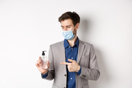 Covid-19, pandemic and business concept. Office worker in medical mask pointing at bottle of hand sanitizer, using antiseptic at work, white background.