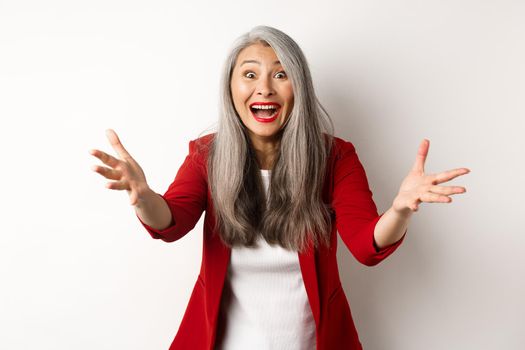 Surprised and friendly asian woman reaching hands forward and smiling, welcoming you, greeting someone, standing over white background.