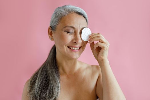 Positive grey haired Asian lady removes makeup from eye with cotton pad posing on pink background in studio. Mature beauty lifestyle