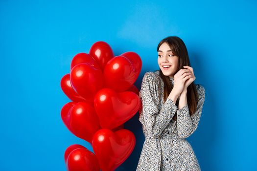 Valentines day. Beautiful romantic girl dreaming off date, standing near lovely heart balloons and smiling, blue background.