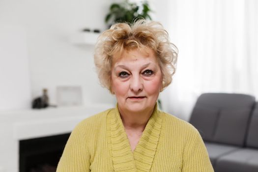 Attractive elderly woman sitting on couch at home, looking at camera, makes video call, communication distantly, modern wireless technologies concept