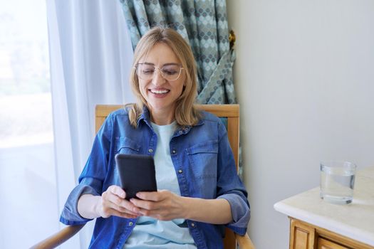 Middle aged woman dressed in casual denim shirt looking at smartphone screen smiling talking, sitting on chair at home. Video call, online communication, work, leisure