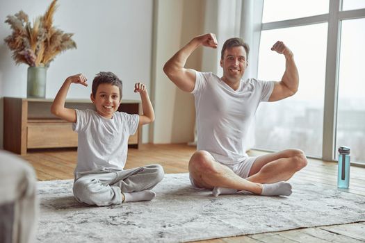 Full length portrait of happy muscular male sitting on mat with son and showing biceps together