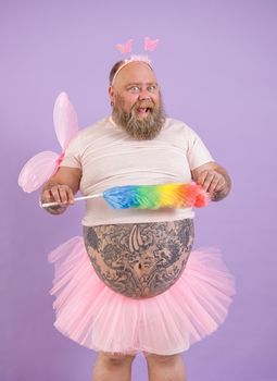 Happy bearded plump man in funny fairy costume with pink wings holds colorful pp duster on purple background in studio