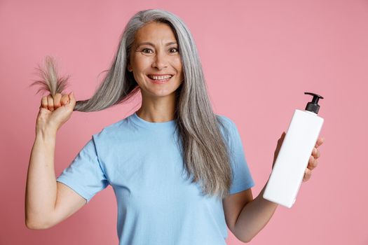 Joyful middle aged Asian woman shows healthy silver hair and blank dispenser bottle of cosmetic products on pink background in studio. Mature beauty lifestyle