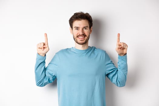 Image of young modern guy with beard showing advertisement on copy space, pointing fingers up and smiling, check it out gesture, white background.