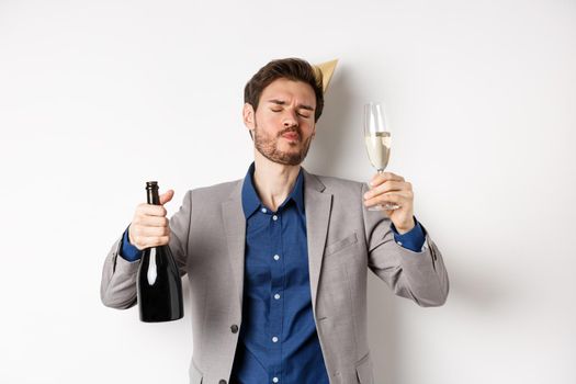 Celebration and holidays concept. Drunk guy having fun at birthday party, dancing with glass of champagne and bottle, wearing suit with cone hat, white background.