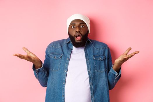Confused and shocked african american man spread hands sideways and drop jaw, staring with awe and amazement at camera, standing over pink background clueless.
