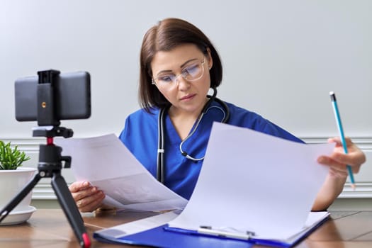 Online doctor consultation, female specialist talking with patient using video call, smartphone on tripod, doctor with stethoscope looking at the phone's webcam
