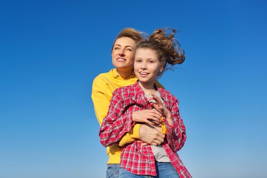 Outdoor portrait of happy smiling mom and child daughter hugging together, blue sky background, family, love, happiness, joy, childhood concept