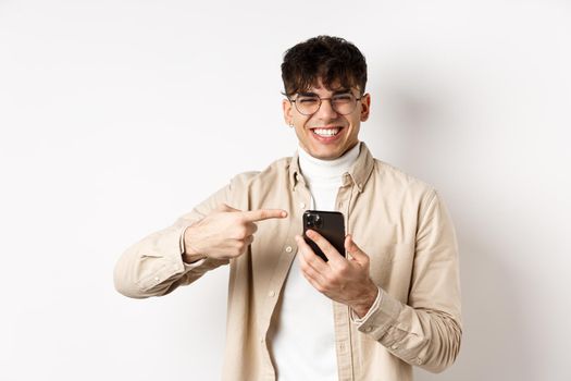 Technology and online shopping concept. Portrait of natural man in glasses laughing and pointing finger at smartphone, showing promo offer on screen, standing white background.