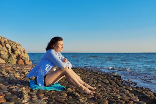 Mature woman sitting on the seashore enjoying the seascape, nature, sunset. Meditating, relaxing, breathing sea air, copy space