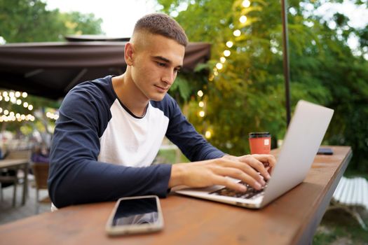 Young man sitting at table and typing on laptop keyboard while working in outdoor cafe, close up