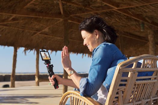 Middle aged woman looking at smartphone webcam talking recording video on sandy beach. Female blogger vlogger using video call on smartphone for blog, online stream, natural sea resort landscape