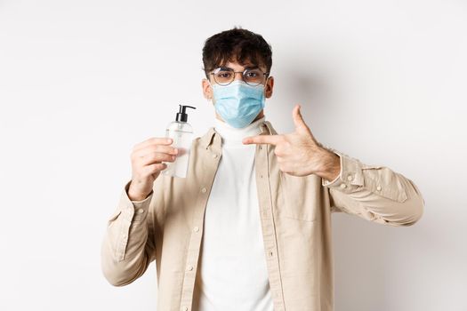 Health, covid and quarantine concept. Cheerful young guy in face mask an glasses pointing finger at bottle of antiseptic, showing good hand sanitizer, white background.