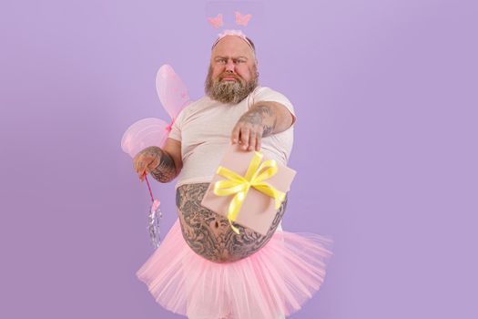 Emotional brutal obese man wearing fairy costume holds magic stick and gift box grimacing on purple background in studio