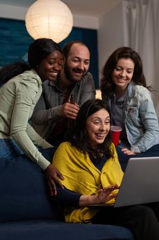 Multi-ethnic friends relaxing on couch laughting while watching fuuny comedy film during movie night at home enjoying spending time together. Group of people hanging out in evening. Friendship concept