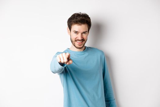 Image of confident young man smiling and pointing at camera, inviting and choosing you, beckon to join him, standing on white background.