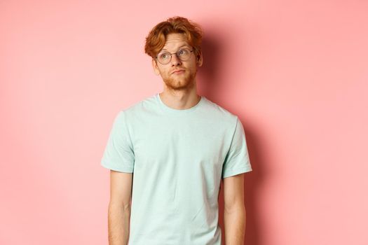 Image of young redhead man with beard, wearing t-shirt and glasses, looking left with bored unbothered face, standing over pink background.