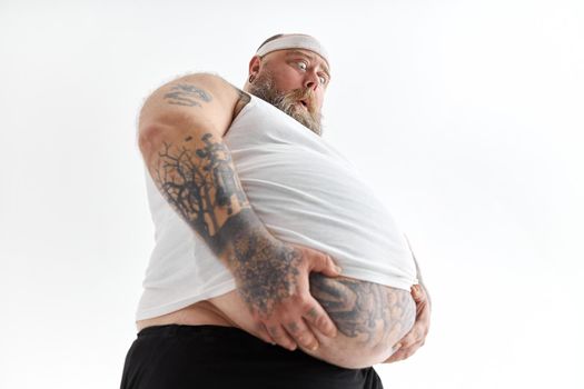 fat man with big belly and tattoes in sports wear is holding his stomach with shocked emotion