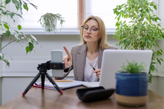 Mature business woman working online from home. Female sitting at table, using smartphone and laptop for remote work, video call, video session, teacher teaching individually remotely