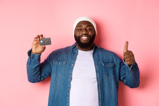 Shopping concept. Handsome african-american man recommending bank, showing credit card and thumbs-up, smiling satisfied, standing over pink background.