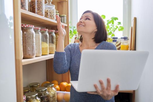 Woman in home kitchen pantry near wooden rack with stored products, holding laptop in hands, looking at screen talking on video communication, food blog, online nutritionist consultation