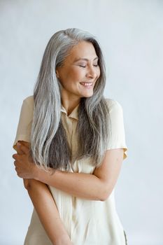 Shying middle aged Asian lady with long silver hair poses on light grey background in studio. Mature beauty lifestyle