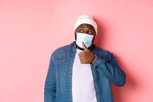 Covid-19, lifestyle and quarantine concept. Cheerful Black man showing thumbs-up and looking left, praising promo offer, approve and like, standing over pink background.