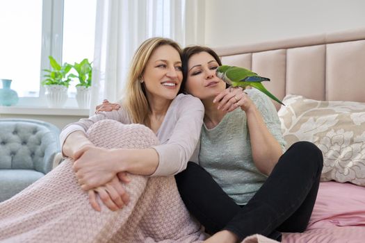 Two happy middle aged women sitting together on the bed with pet green parrot. Same sex female couple, relationship, lifestyle, people concept