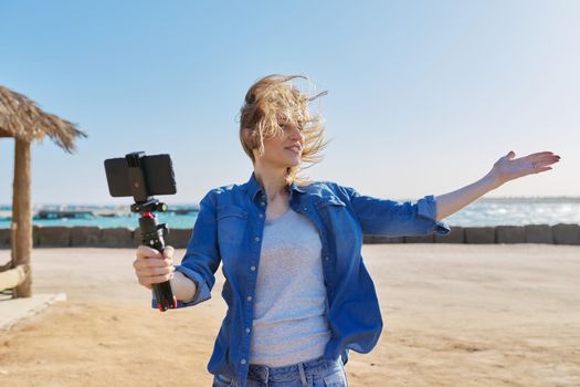 Middle aged beautiful woman recording video on smartphone, sunset sea beach background. Female blogger, vlogger, talking to followers using video call, recording stream