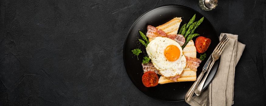 Breakfast or lunch with Fried egg, bread toast, green asparagus, tomatoes and bacon on black plate. Top view. Copy space. Banner.