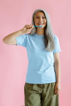 Positive long haired mature Asian model in blue t-shirt holds toothbrush on pink background in studio. Oral cavity hygiene