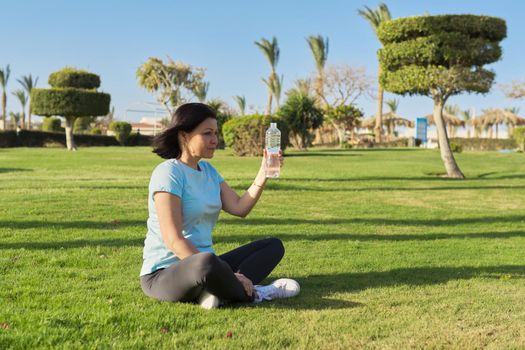 Mature woman doing morning exercises on green grass in park, with a bottle of water, sports, healthy active lifestyle of middle-aged people