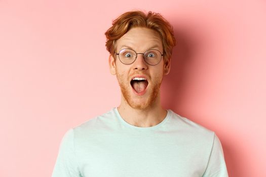 Close up of surprised redhead man raising eyebrows and scream with joy, checking out cool promo, looking amazed at camera, standing over pink background.