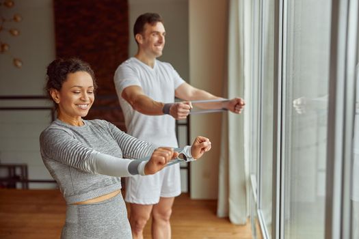 Jolly man and woman are standing before window and training arms with resistance bands