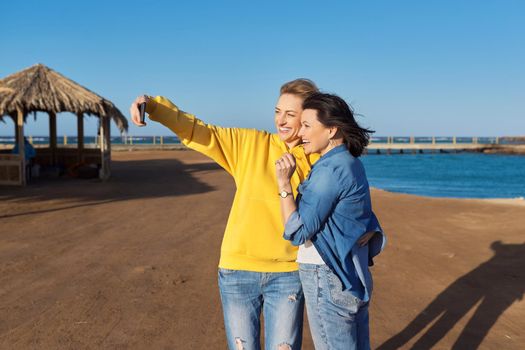 Two mature happy women taking selfie photo together using smartphone. Sea beach, resort, nature background. Friendship, family, same-sex couple, vacation, weekend, relaxation at sea concept