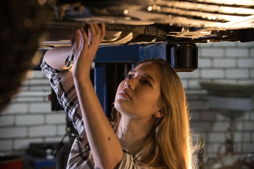 Car repair service. Young sexy mechanic woman standing under the car holding a wrench and looking up. Mid shot