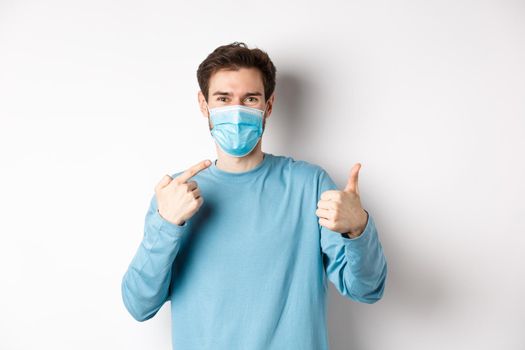 Coronavirus, health and quarantine concept. Smiling man pointing at his medical mask and showing thumb up, standing on white background.