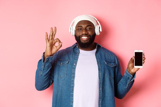 Happy hipster guy listening music on headphones and showing mobile screen, smiling satisfied, showing okay sign in approval, like playlist, standing over pink background.