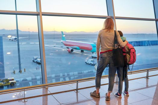 Airport passengers, standing with their backs family mother and daughter child looking at planes in panoramic window, awaiting boarding, copy space