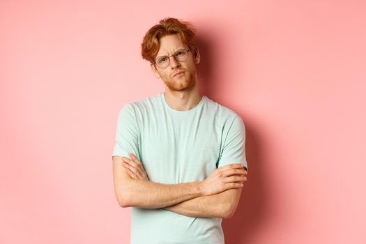 Young man with red hair and beard, wearing glasses and t-shirt, cross arms on chest, frowning while staring with skeptical and doubtful expression, standing over pink background.