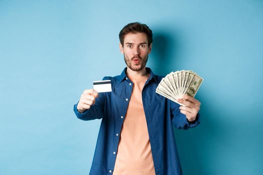 Excited modern guy showing money and plastic credit card, saying wow amazed, standing on blue background.