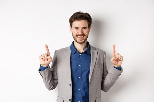 Confident successful businessman pointing fingers up, smiling and showing company logo, standing in suit on white background.