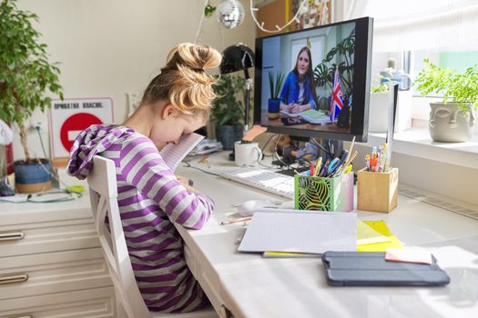 E-education, online English lesson. Girl child sitting at table at home looking at computer monitor, talking learning language with young teacher