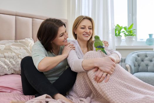 Two happy middle aged women sitting together on the bed with pet green parrot. Same sex female couple, relationship, lifestyle, people concept